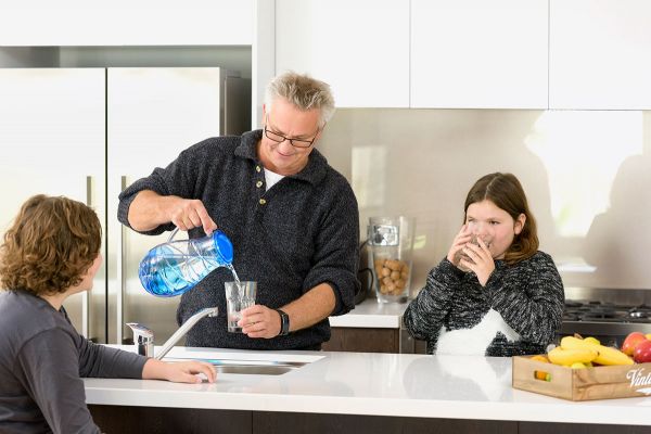 A middle-aged man pouring a glass of water from a jug, over a sink. A young girl stands to his left, drinking a glass of water. A young boy is sitting at the kitchen counter, looking at the man.